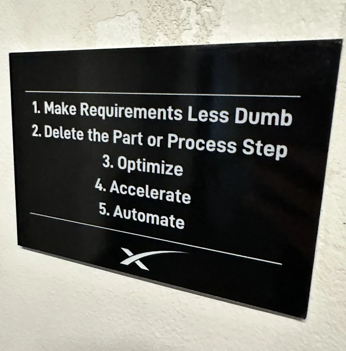 A black sign on a workshop wall listing the five steps: make requirements less dumb, delete the part or process step, optimize, accelerate, automate.